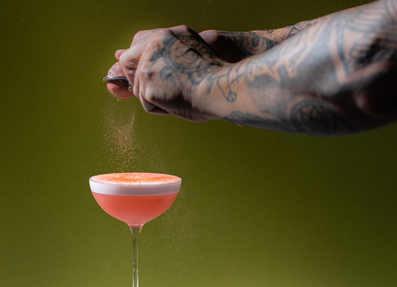 Image of a mixologist with tattooed arms sprinkling peach dust on a peach melba sour cocktail.